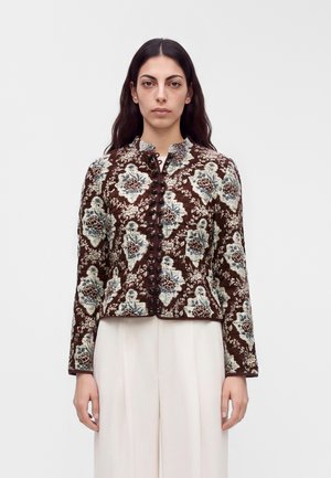Une femme porte une veste marron à motif floral avec des boutons foncés et un pantalon blanc sur un fond clair uni.