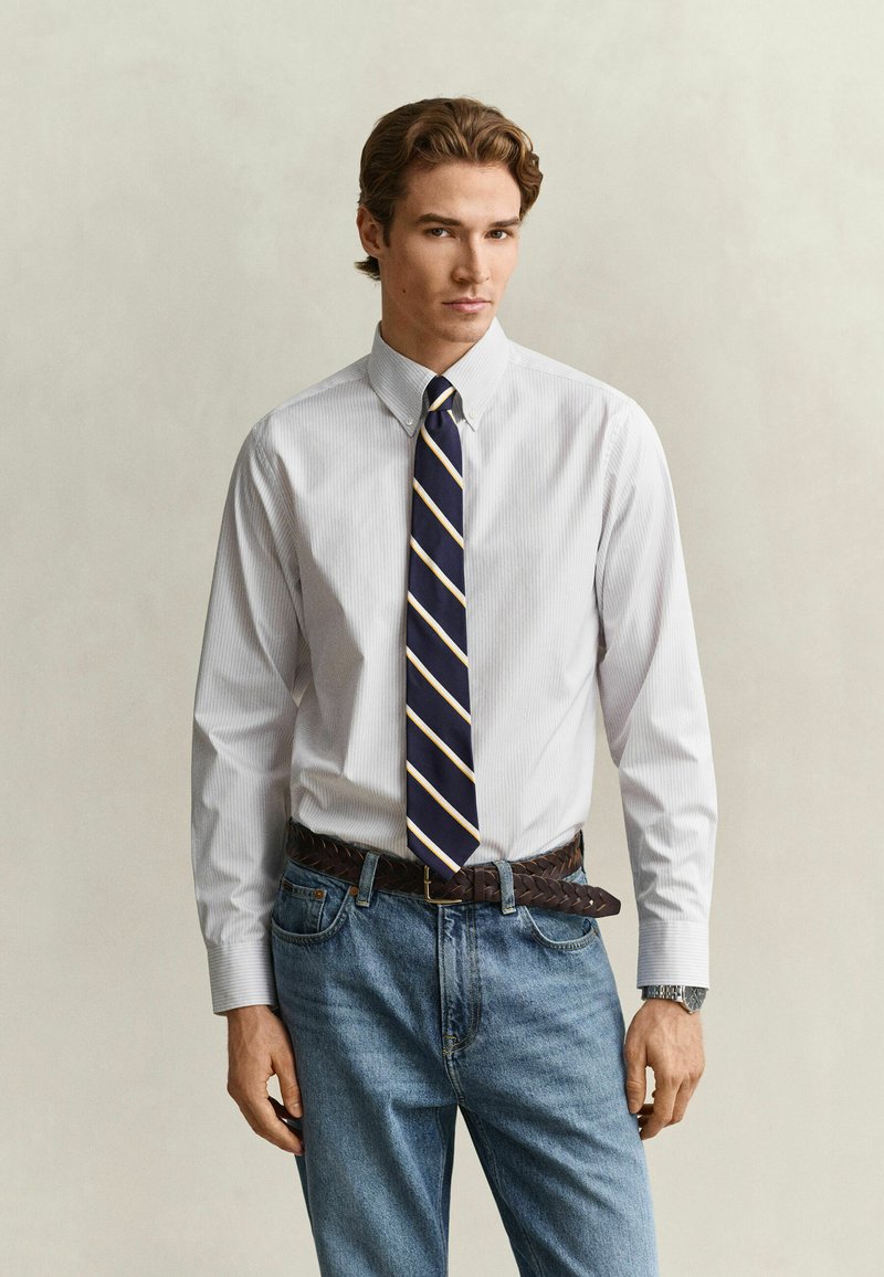 Young man with light brown hair wearing a white button-up shirt, navy blue striped tie, braided brown belt, and blue jeans.