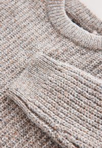 Pull en tricot beige et gris avec poignets et col côtelés, montrant la texture détaillée du tissu en laine.