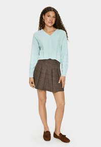 Ung kvinna med lockigt hår som bär en ljusblå kabelstickad tröja, en brun veckad minikjol och bruna loafers, stående framför en vit bakgrund.