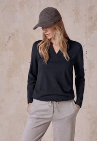 Svart långärmad topp med V-ringning och texturerad krage, kombinerad med ljusgrå byxor med dragsko och en mörk stickad mössa.