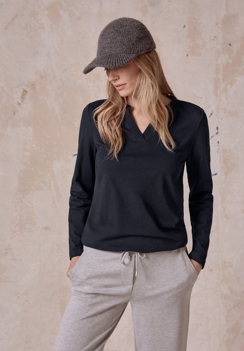 Svart långärmad topp med V-ringning och texturerad krage, kombinerad med ljusgrå byxor med dragsko och en mörk stickad mössa.