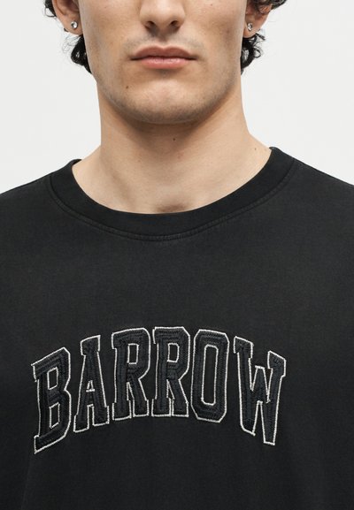 Κοντινό πλάνο ενός ατόμου που φοράει μαύρο μπλουζάκι με στρογγυλή λαιμόκοψη και κεντημένο το "BARROW" με μεγάλες λευκές επισήμανσεις στο στήθος.