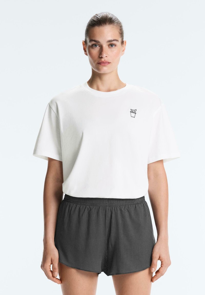 Femme aux cheveux tirés en arrière portant un t-shirt blanc uni avec un petit motif de plante et un short noir à carreaux, debout devant un fond blanc.