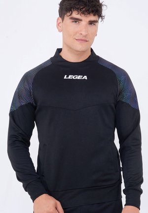 Uomo che indossa una felpa sportiva nera LEGEA con pannelli geometrici sulle spalle, in piedi su sfondo bianco uniforme.