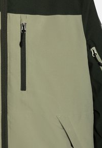 Urban jacket featuring a color-block design in dark green and olive green, with zippers, textured fabric, and angled pockets.