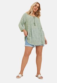 Ljusgrön blus med blommönster, rundad halsringning, knyt-detalj och 3/4-ärmar, tillsammans med ljusa denimshorts och vita sandaler.