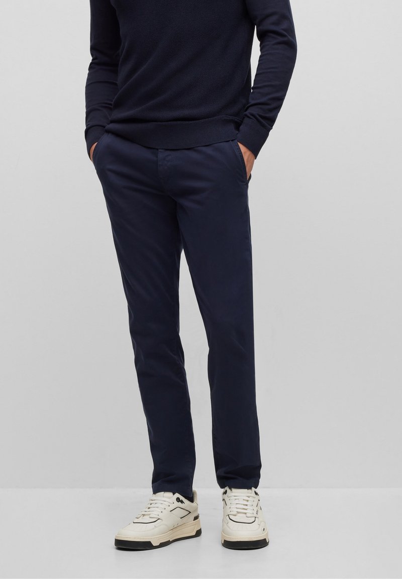 Homme portant un pull bleu marine et un pantalon ajusté assorti avec des baskets blanches, debout les mains dans les poches devant un fond uni.