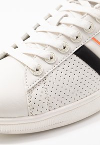 Zapatilla de cuero blanca con diseño perforado, cordones planos blancos, ojales plateados, detalles de franjas en naranja y negro, y suela de goma texturizada.