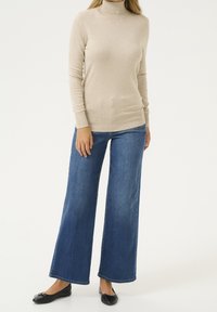 Pull col roulé beige en maille avec des poignets côtelés, associé à un jean en denim bleu à jambes larges. Des ballerines noires complètent la tenue.