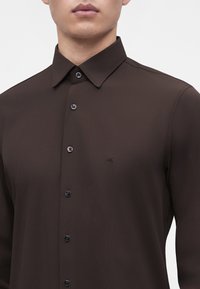 Homme portant une chemise marron foncé à boutons avec un col pointu et un petit logo discret sur la poitrine gauche sur un fond uni.