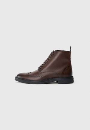 Bruine leren enkellaarzen met een gladde afwerking, ronde neus en vetersluiting. Beschikt over decoratieve brogue-details en een zwarte rubberen zool.