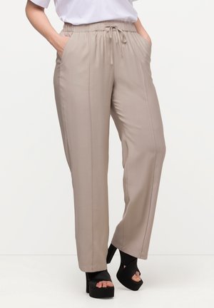 Person trägt beige Kordelhosen und schwarze Sandaletten mit hohen Absätzen, Hände in den Taschen, steht vor einem einfarbigen Hintergrund.