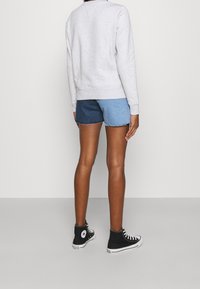Persona che indossa una felpa grigia chiara, pantaloni shorts in denim bicolore (blu scuro e blu chiaro) e sneakers nere alte, in piedi contro un muro bianco.