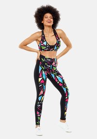 Schwarzes Sportoutfit, das einen Sport-BH mit bunten geometrischen Mustern und hochgeschnittene Leggings mit ähnlichen Designs umfasst. Weiße Turnschuhe.
