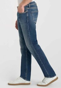 Jean en denim de couleur bleu foncé avec une coupe droite, un design à cinq poches et des coutures visibles ; associés à des baskets blanches.