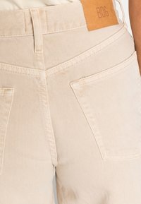 As calças de ganga bege apresentam uma etiqueta de pele com "BDG". O design inclui dois bolsos traseiros com costura visível e um corte reto.