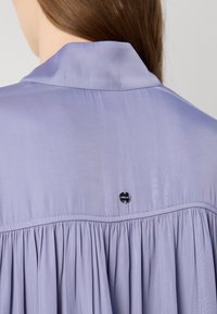 Levandulová blůza s límečkem, s nařasenou strukturou a malým černým logem na knoflíku na zádech. Měkká, hladká tkanina.