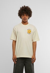 T-shirt à manches courtes de couleur crème avec un col rond, arborant un motif de visage souriant jaune et un texte noir sur la poitrine gauche.