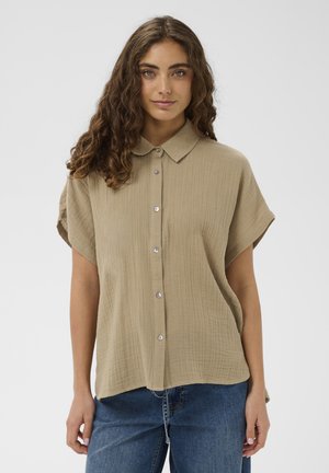 Mladá žena s dlhými kučeravými hnedými vlasmi, oblečená v voľnej béžovej košeli na gombíky s krátkym rukávom a modrých džínsoch, stojaca na bielom pozadí.