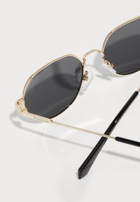 Gold metal sunglasses with oval dark lenses, featuring clear nose pads and black temple tips. Subtle branding on the frame.