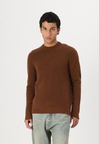 Pulover din lână maro cu guler rotund, mâneci lungi și manșete îndoite, asortat cu blugi din denim de un albastru deschis. Textură moale, design minim.