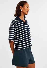 Navy ja valkoinen raidallinen polo-paita, jossa on röyhelöinen rakenne ja lyhyet hihat, yhdistetty pimeisiin sinisiin laskostettuihin shortseihin.