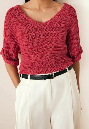 Kobieta nosząca luźny, czerwony sweter z podwiniętymi rękawami, włożony do białych spodni z wysokim stanem i czarnym paskiem.