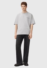 T-shirt gris à manches courtes avec un col rond et un logo brodé ; associé à un jean noir à jambe large et des bottines noires.