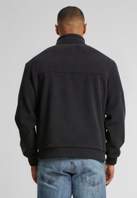 Man som bär en svart fleecejacka och blå jeans, står med ryggen vänd mot kameran framför en enfärgad grå bakgrund.