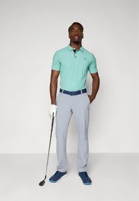 Polo verde claro com três botões, calças cinzentas, cinto azul-marinho, luva branca de golfe e sapatos de golfe azuis; segurando um taco de golfe.