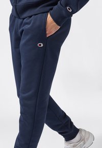 Persona che indossa pantaloni tute Champion blu navy, con una mano in tasca e sneakers bianche su uno sfondo chiaro e semplice.