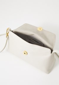 Bílá kožená crossbody taška s hladkým povrchem, zlatým kováním a jedním magnetickým uzávěrem; interiér má sloty na karty a textilní podšívku.