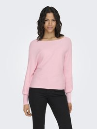 Donna con capelli scuri ondulati, indossa un maglione rosa chiaro a coste con maniche a palloncino e pantaloni neri aderenti, in piedi davanti a uno sfondo bianco semplice.