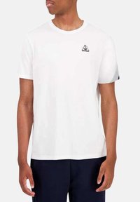 T-shirt bianco a maniche corte in cotone, con un piccolo logo grafico nero sul lato sinistro del petto e un colletto rotondo.