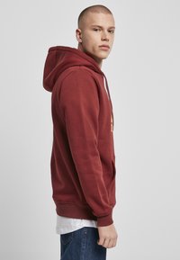 Sudadera color burdeos con un bolsillo frontal, capucha ajustable con cordón y puños de canalé. Superpuesta sobre una camiseta blanca con un detalle de logo.