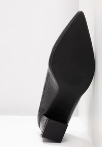 Chaussure en cuir noir texturé à bout pointu reposant sur son talon, montrant une semelle détaillée et un talon bloc sur fond blanc.