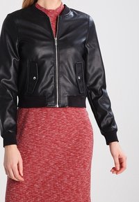 Černá kožená bomber bunda se zapínáním na zip, dvěma předními kapsami, žebrovanými manžetami a lemem, nošená přes červené texturované šaty.