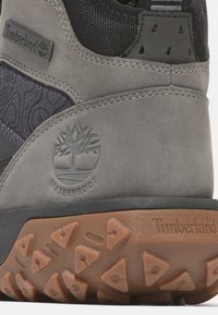 Szare wodoodporne buty Timberland mają teksturowaną czarną tkaninę na wierzchu, wyraźne logo i solidną brązową podeszwę z trójkątnymi wypustkami zapewniającymi przyczepność.