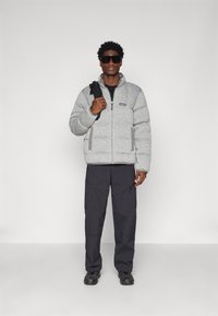 Grå pufferjacka med dragkedja, sidofickor och texturerad yta. Matchad med svarta byxor och mörka skor. Modellen bär solglasögon och håller en väska.