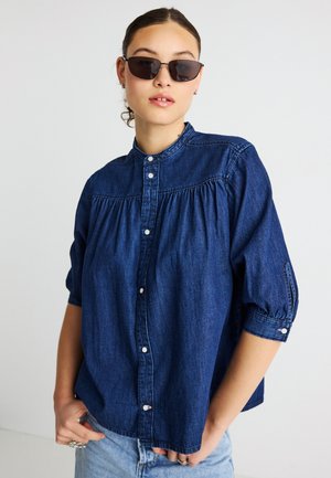 Donkerblauw denim overhemd met een mandarijnen kraag, knoopsluiting aan de voorkant, geplooide details en opgerolde mouwen, gecombineerd met zwarte zonnebrillen.