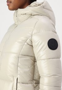 Beige pufferjack met een glanzende, gequiltte buitenkant en een capuchon. Heeft een zwart, rond logo-patch op de linker mouw. Voorzien van een ritssluiting aan de voorkant.