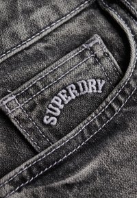 Grijze denim stof met zichtbare stiksels. Een stoffen label dat in de zak is genaaid, draagt de tekst "SUPERDRY" in witte geborduurde letters.