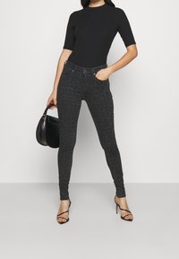Los jeans de estampado de leopardo negro presentan un ajuste slim, una cintura de altura media y una textura sutil. Combinados con una blusa negra con canalé y tacones con tiras.