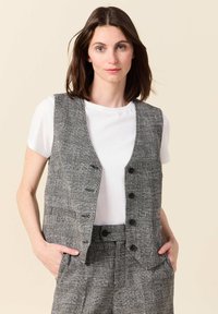 Grauer karierten ärmellose Weste mit vier Knöpfen, Seitentaschen und einem hellen T-Shirt darunter. Glatte Textur, taillierte Passform.