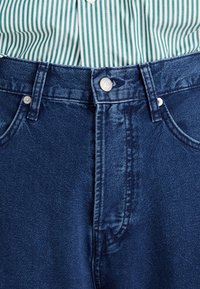 Jeans iz denima v temno modri barvi s klasičnim krojem, ki imajo zapiranje na srebrn gumb in bež šive, v kombinaciji z zeleno in belo črtasto srajco.