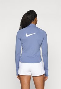 Langärmliges Sportoberteil in Hellblau, aus glattem Material, mit hohem Kragen und weißem Nike-Logo auf der Rückseite, kombiniert mit weißen Shorts.