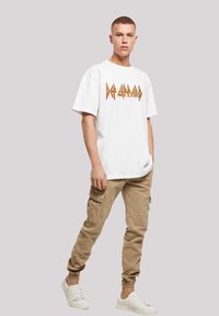 Weißes oversized T-Shirt mit orangem und gelbem "Def Leppard"-Logo, kombiniert mit khakifarbenen Cargo-Hosen und weißen Sneakern. Glatte Textur, lässige Passform.