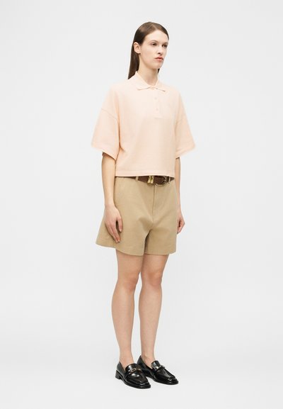 Γυναίκα που φοράει ροζ ανοιχτό ροζ κοντομάνικο πόλο πουκάμισο, μπεζ σορτς με ζώνη και μαύρα loafers, στέκεται μπροστά σε λευκό φόντο.