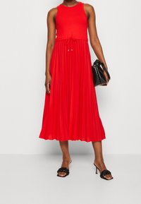 Rode mouwloze jurk met een geribde taille, een getailleerde taille en een vloeiende geplooide rok. Draag met een zwarte handtas en sandalen met hak.